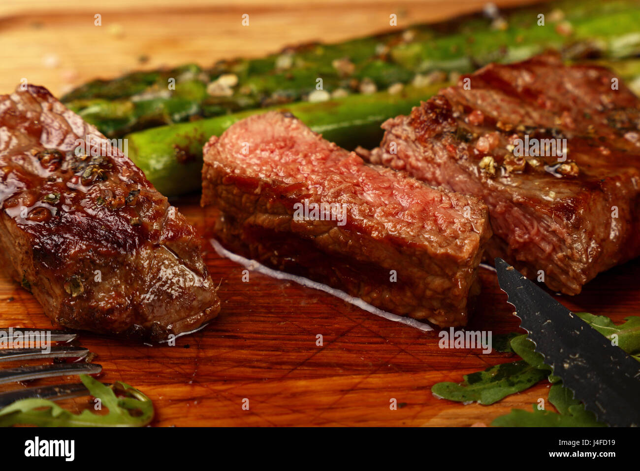 Cut slices of grilled juicy medium cooked beefsteak served on wooden board with fresh green rocket salad, fork and knife, close up, high angle view Stock Photo