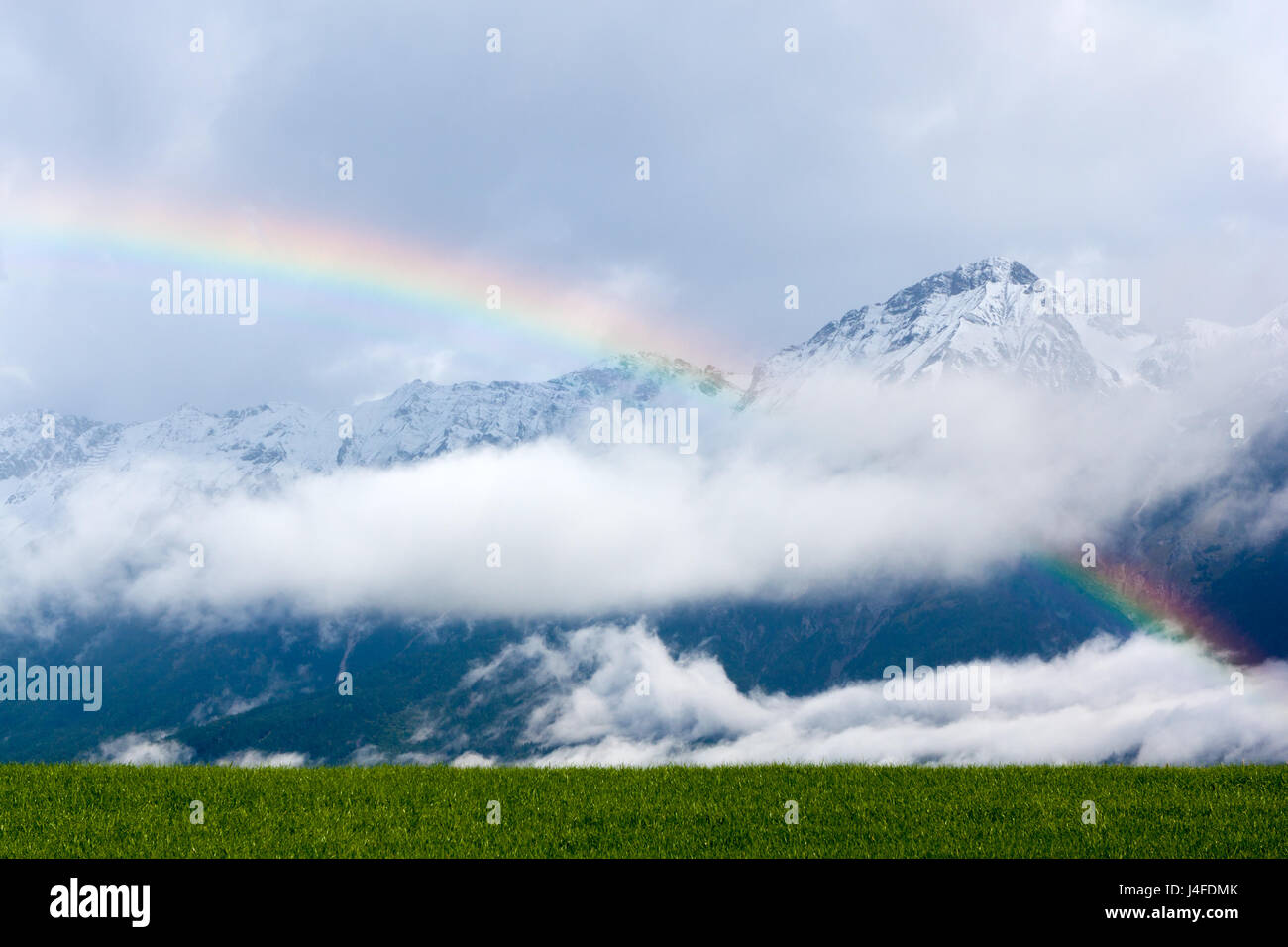 Rainbow over a green meadow in the Alps, with snowcapped hills in the background. Stock Photo
