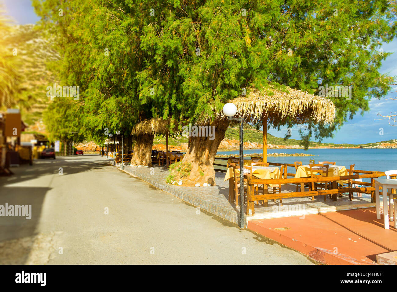 Tables with thatched umbrellas are on the road at beach cafe on the seafront. Livadi beach in sea bay of resort village Bali in may. Bali, Crete, Gree Stock Photo