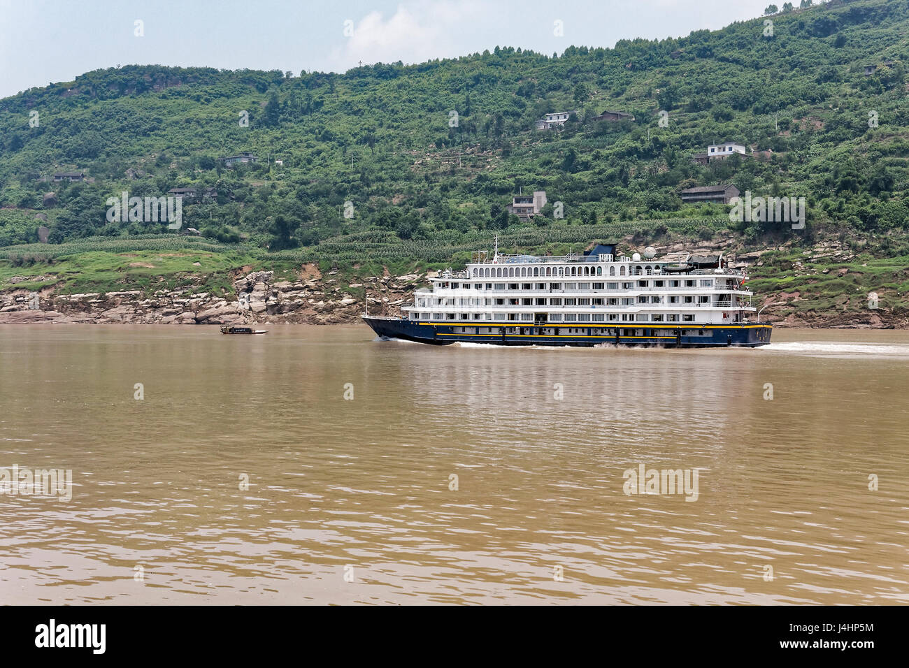 On the Yangtze River a cruise ship sails upstream past a small town nestled on steep wooded slopes. Stock Photo