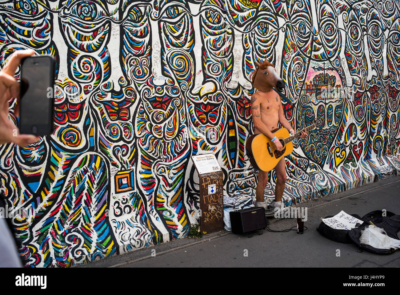 A street busker wearing little else than a horse head mask and a guitar as he sings in front of a mural at Berlin's East Side Gallery. Stock Photo