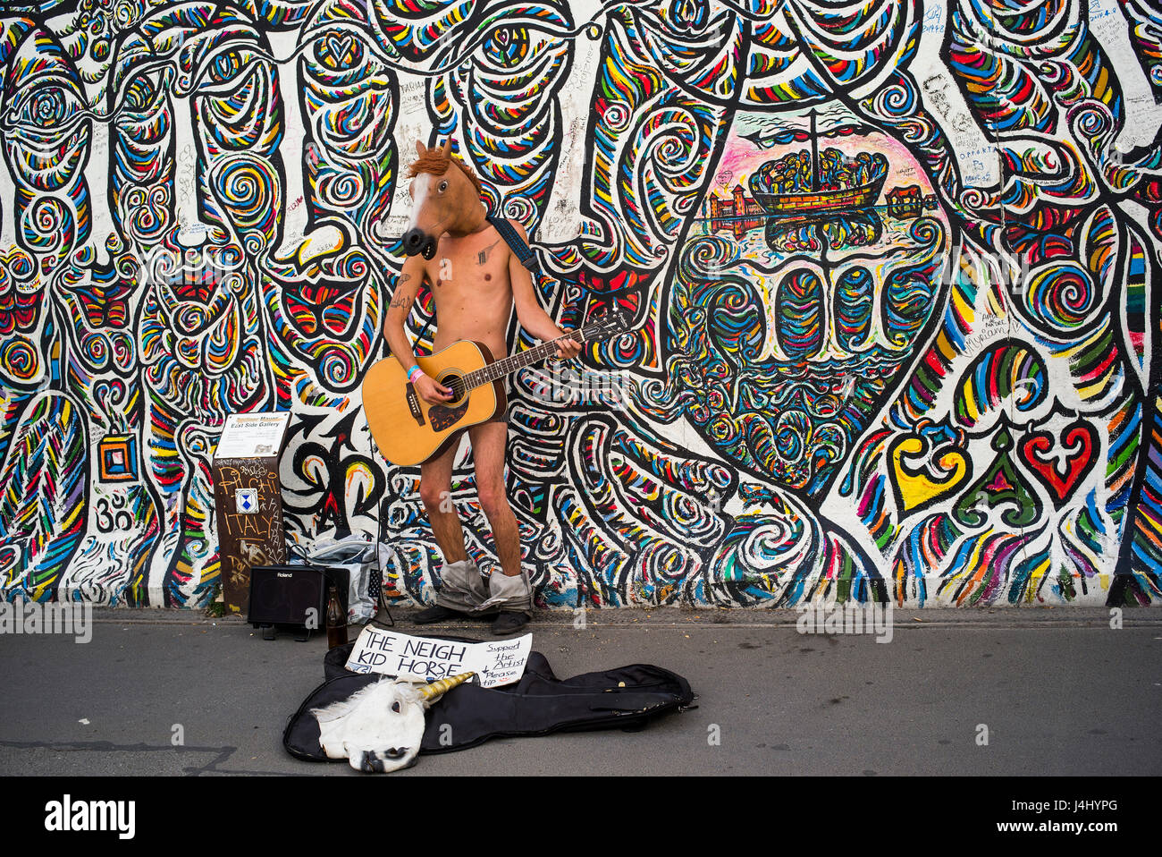 A street busker wearing little else than a horse head mask and a guitar as he sings in front of a mural at Berlin's East Side Gallery. Stock Photo