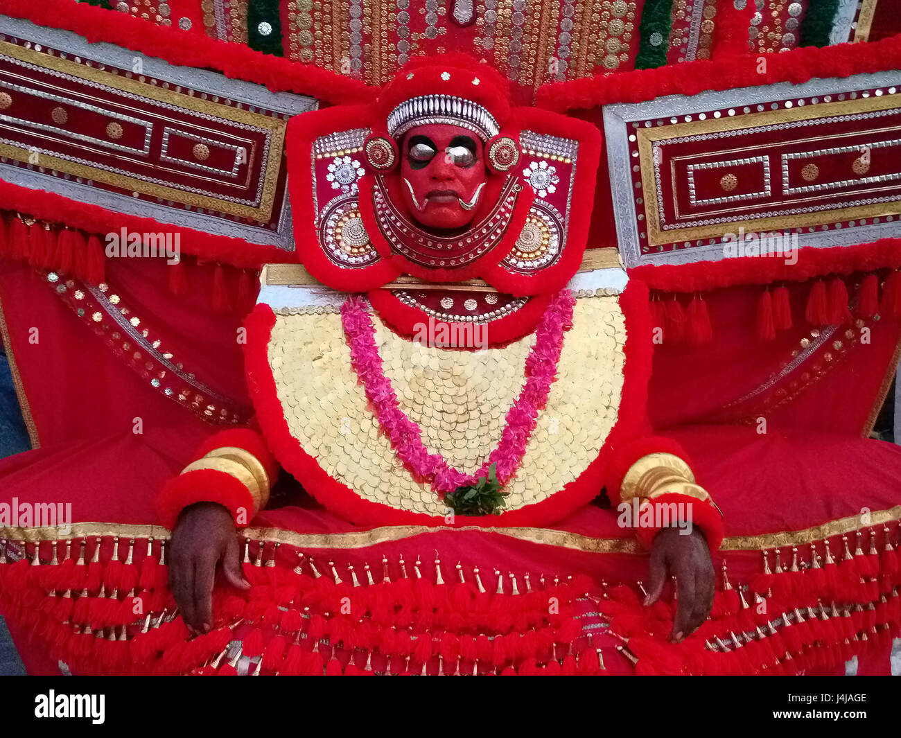 Artist of the ancient art theater Kathakali in suit and mask: bright red wide clothing, garland of red flowers, red mask with metal eye sockets, Keral Stock Photo