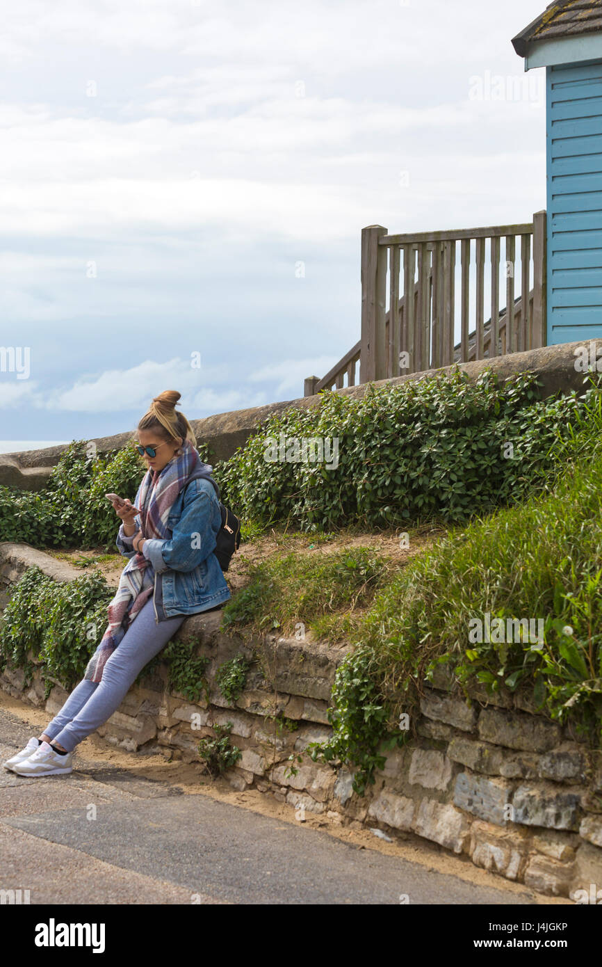 Young lady sitting on wall looking at mobile phone by beach huts at Alum Chine, Bournemouth in May Stock Photo