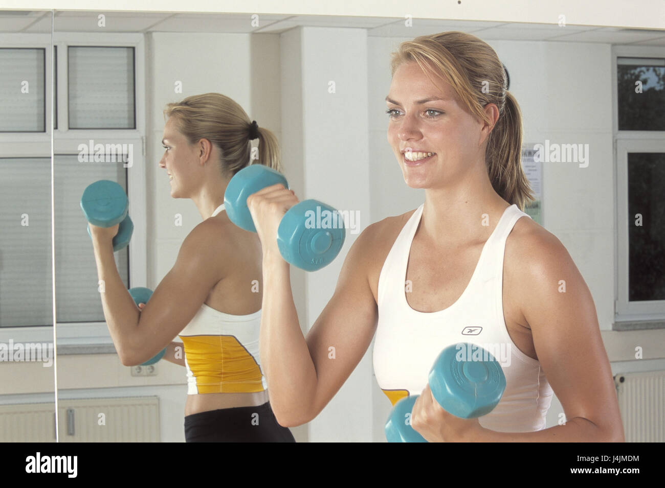 Fitness studio, woman, young, stand, tighten dumbbells, fitness practise, at the side, half portrait fitness room, sportswoman, blond, sports clothes, sportily, sport, hobby, leisure time, fitness, health, motion, practise, musculature, muscles, arms, biceps, arm musculature, to arm muscles, strain, muscle construction, training, train, dumbbell training, weights, short dumbbells, position, position, smile, inside, background, reflector, reflexion Stock Photo