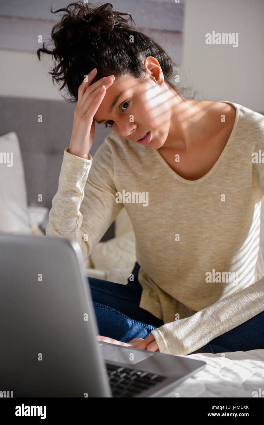 Frustrated Hispanic woman sitting on bed using laptop Stock Photo