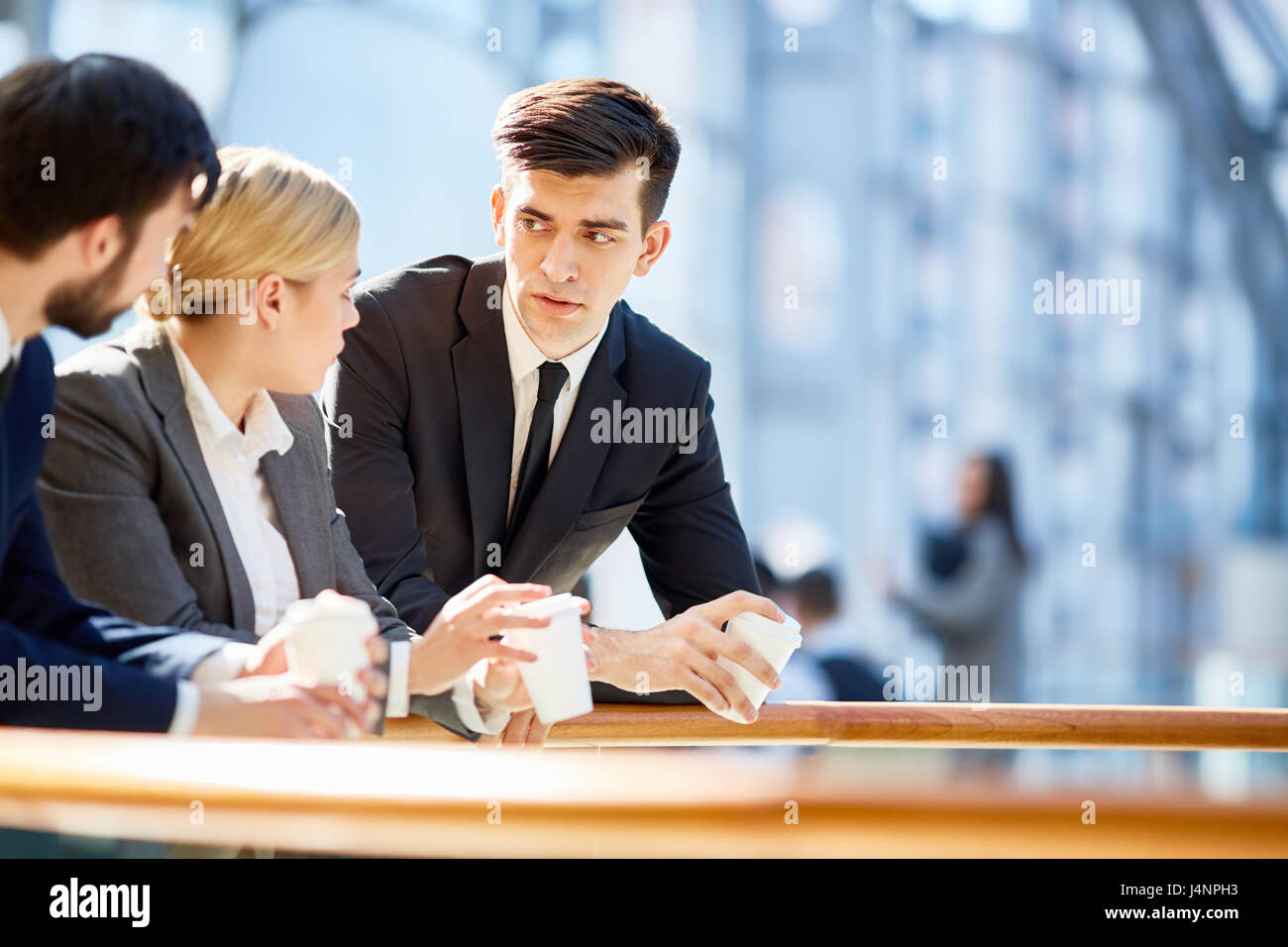Portrait of three business people, one woman and two men, discussing work at break  in modern office building leaning on railing and holding coffee cu Stock Photo