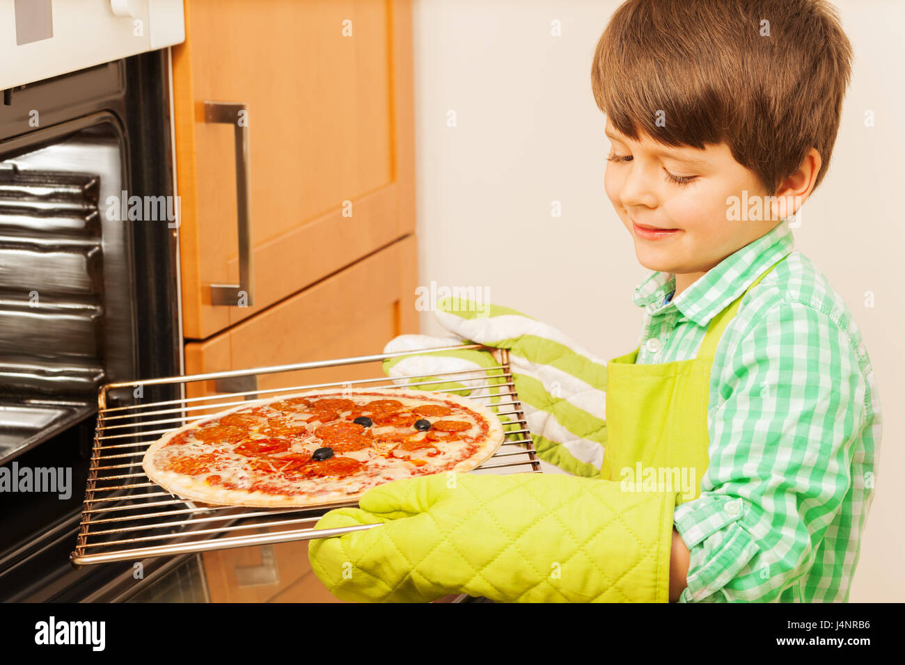 Little cook, six years old boy in mittens holding tray with tasty pizza, standing next to the oven Stock Photo