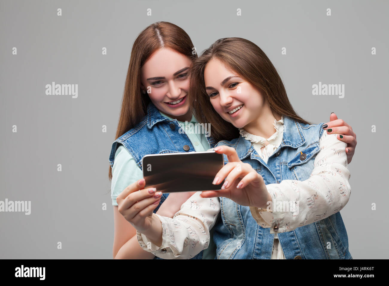 Two smiling girlfriends in denum jackets makes selfie on phone camera in studio. Female friendship. Leisure of happy girls Stock Photo