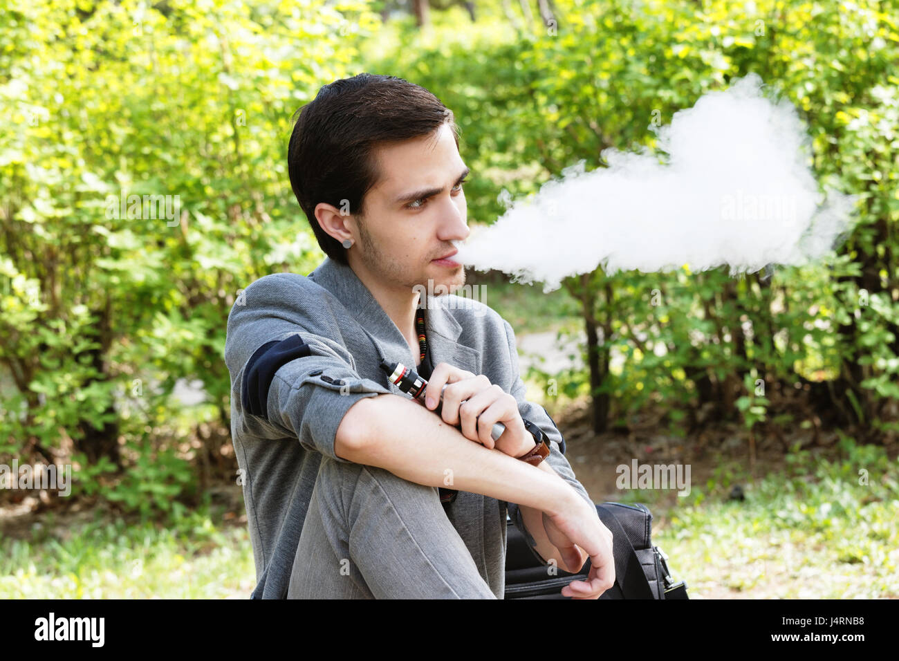 Thoughtful young businessman smokes a vape in a park in the spring afternoon Stock Photo