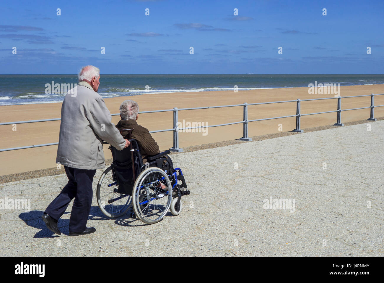 Retired husband taking disabled elderly wife in wheelchair for a walk on promenade along the coast on a cold sunny day in spring Stock Photo