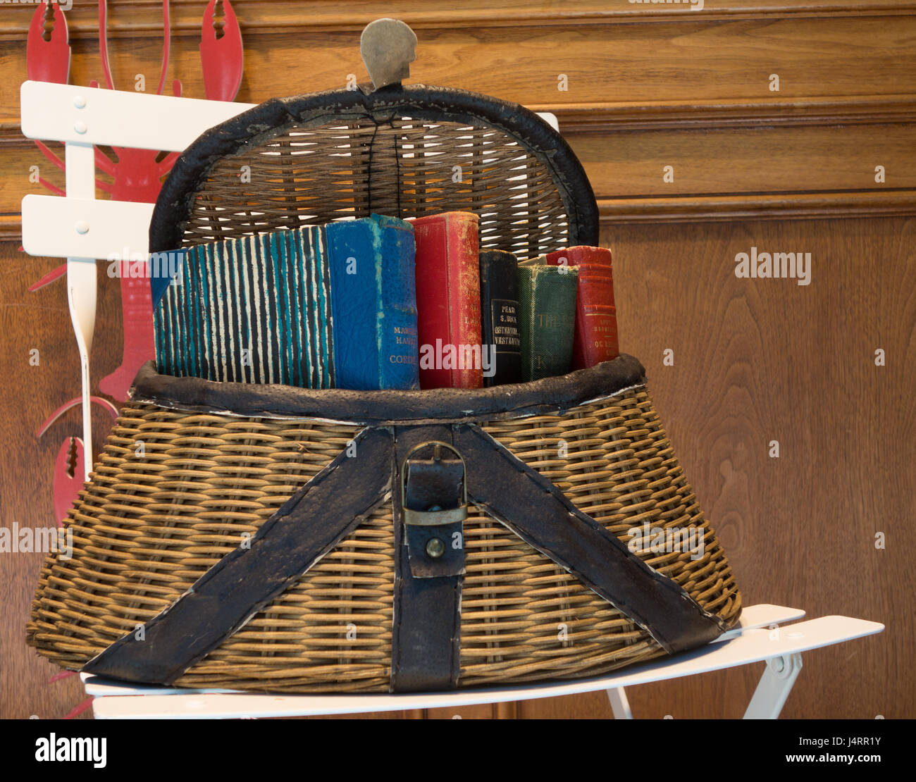 A still life featuring a woven basket with leather trim containing a variety of old books, on a white chair Stock Photo