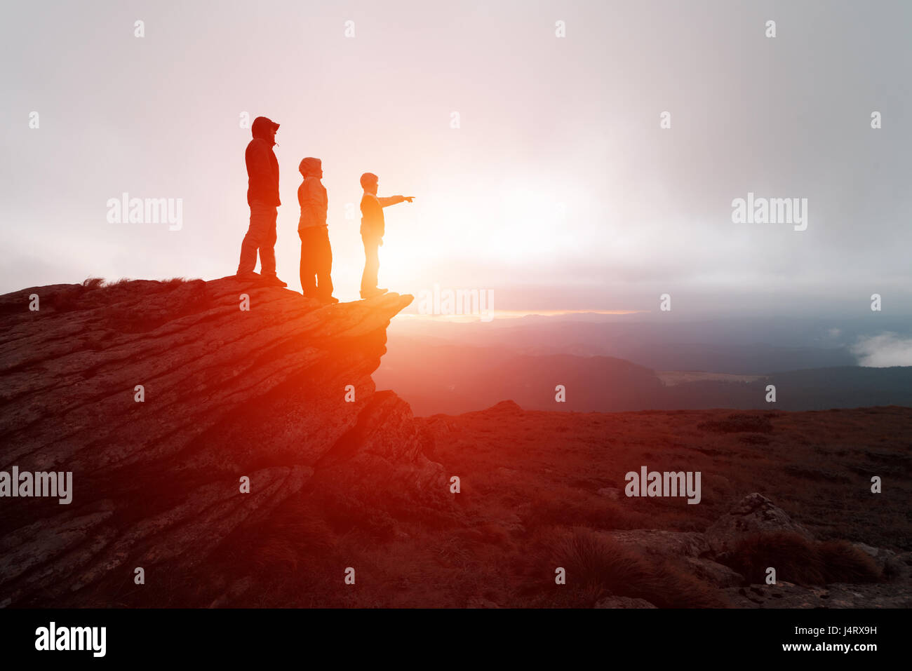 Family of tourists staying on the edge of the cliff against the backdrop of an incredible mountain landscape. Evening time and orange sunset Stock Photo