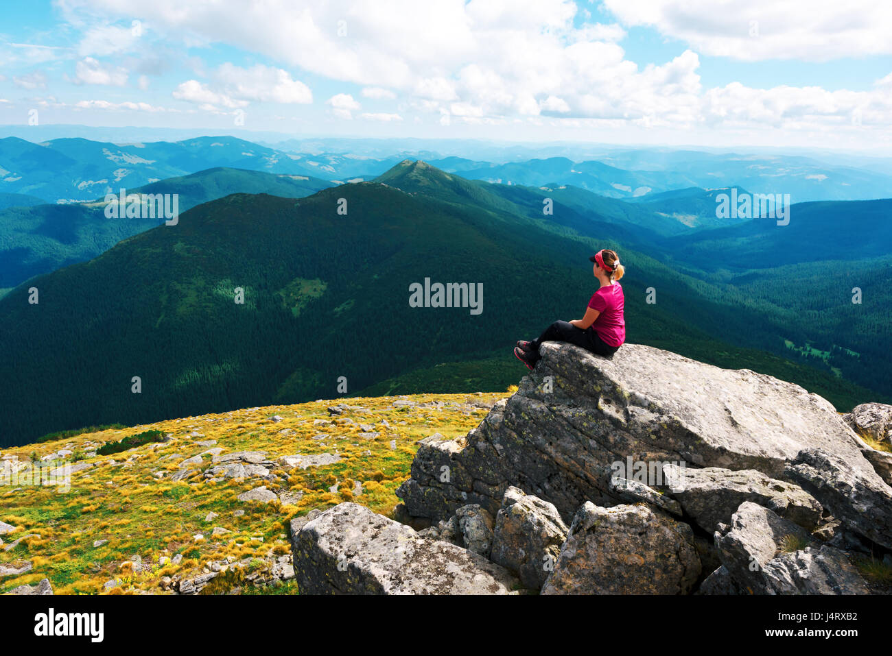 A lone tourist sitting on the edge of the cliff against the backdrop of an incredible mountain landscape. Sunny day and blue sky Stock Photo