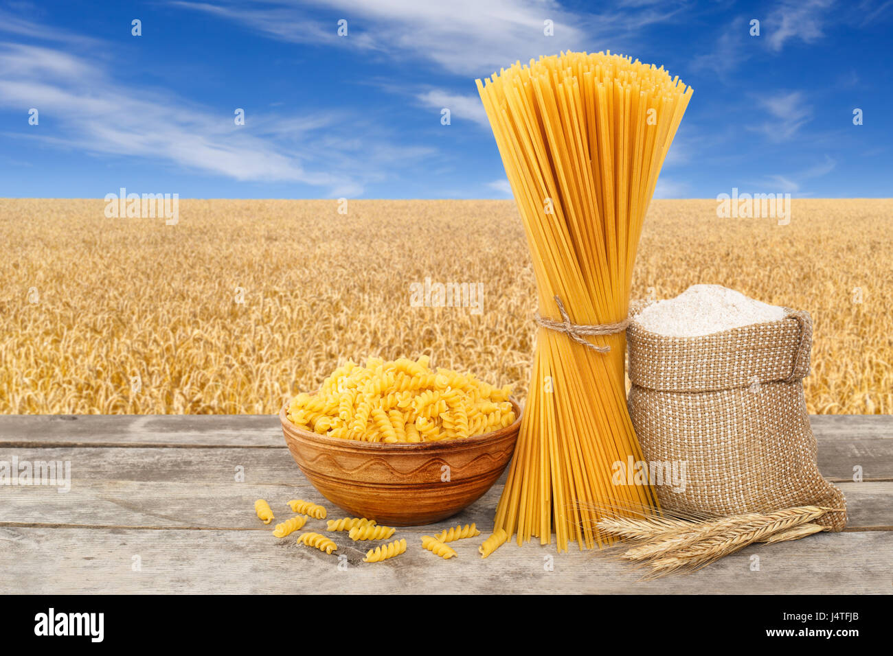 uncooked pasta, spiral shaped and spaghetti, wholegrain flour in bag on table with ripe cereal field on the background. Golden wheat field with blue s Stock Photo