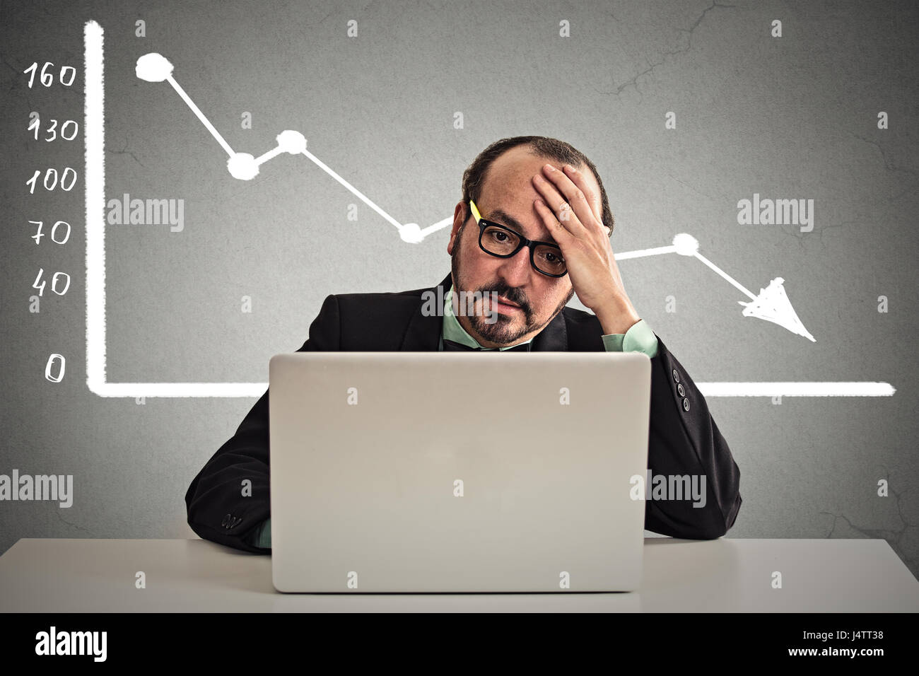 Frustrated stressed business man sitting at table in front of computer with financial market chart graphic going down on grey office wall background.  Stock Photo