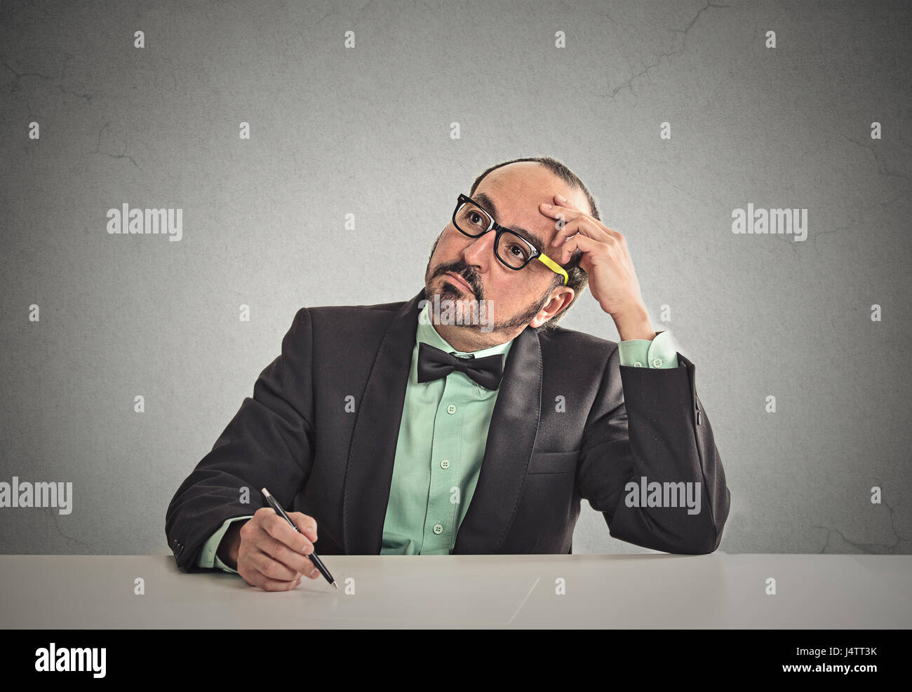 Confused teacher sitting at table scratching his head thinking what idea to write isolated office grey wall background with copy space. Human face exp Stock Photo