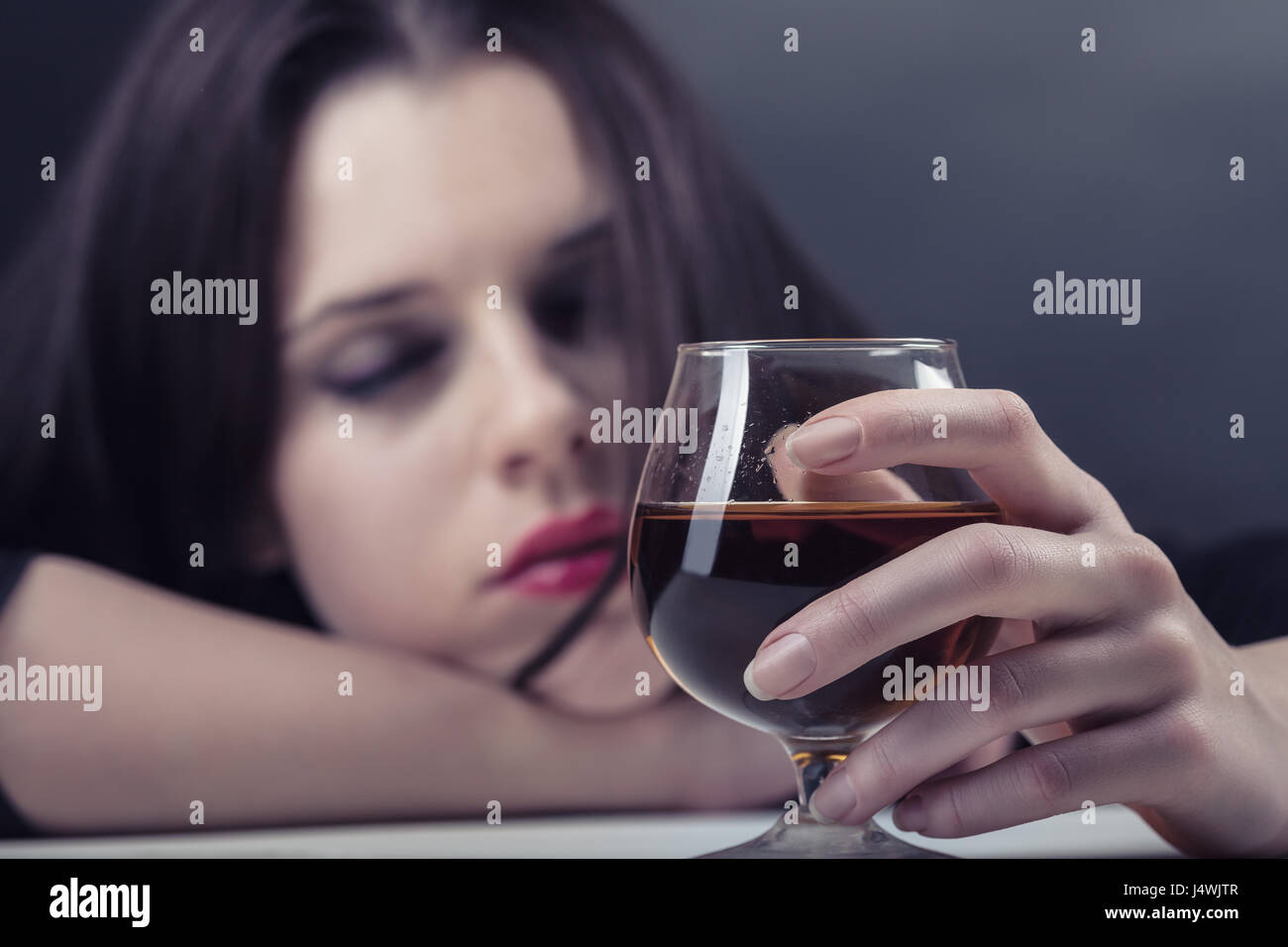 Young beautiful woman in depression, drinking alcohol on dark background. Focus on the glass Stock Photo