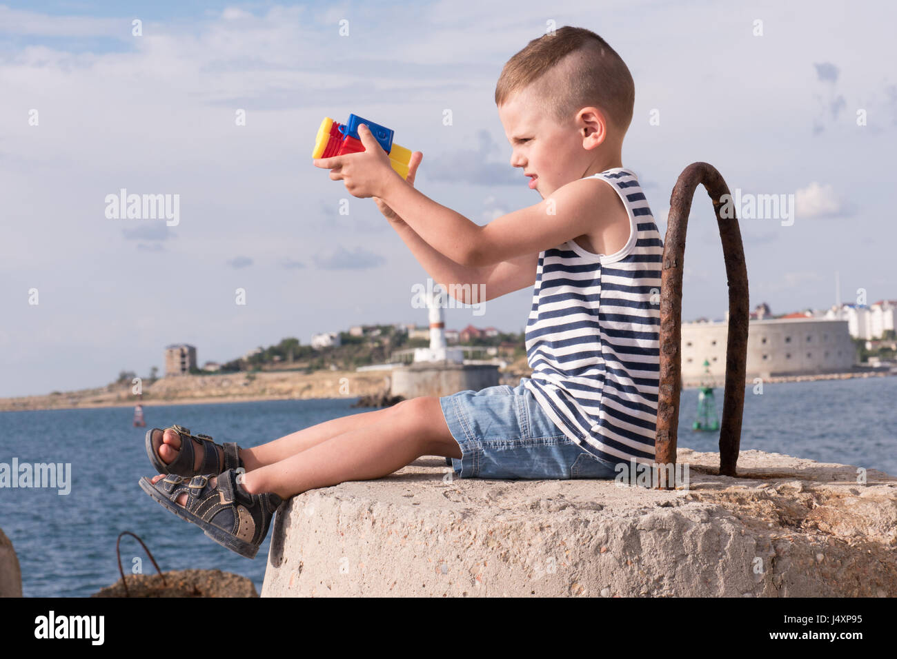 cute small boy in the vest sitting on the breakwater with binoculars on