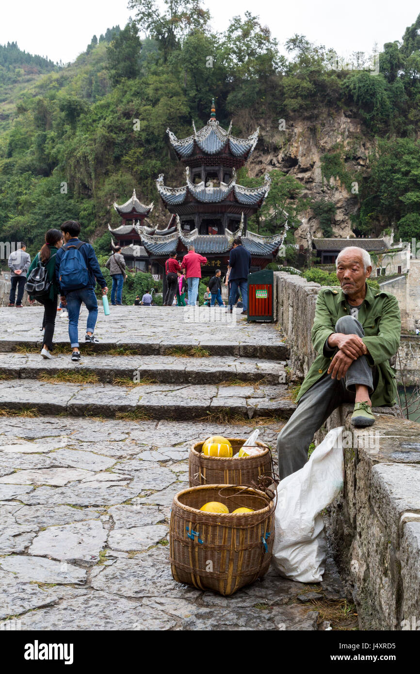 Zhenyuan, Guizhou, China.  Pomelo Vendor Sitting on Wall of Zhusheng Bridge across the Wuyang River. Stock Photo