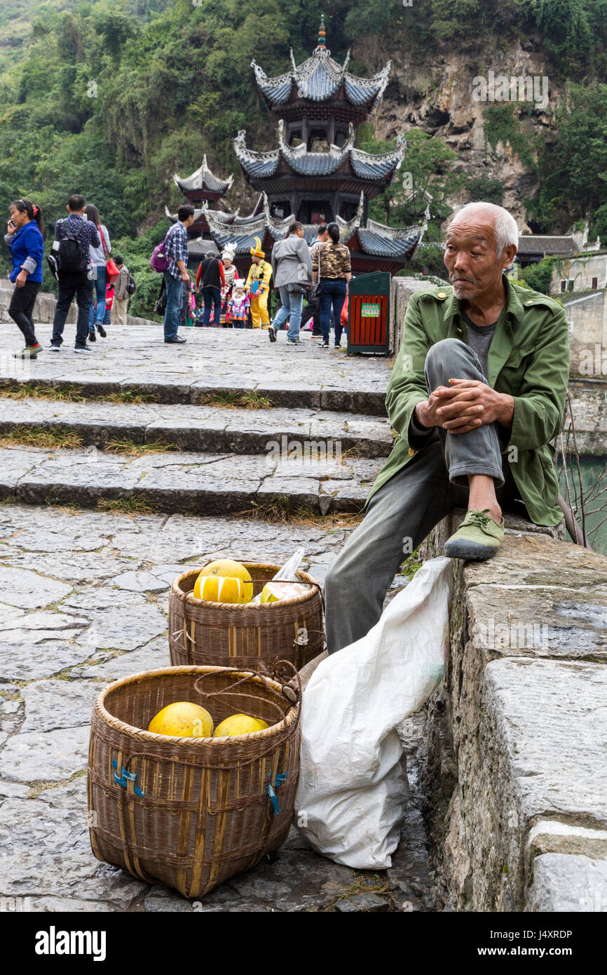 Zhenyuan, Guizhou, China.  Pomelo Vendor Sitting on Wall of Zhusheng Bridge across the Wuyang River. Stock Photo