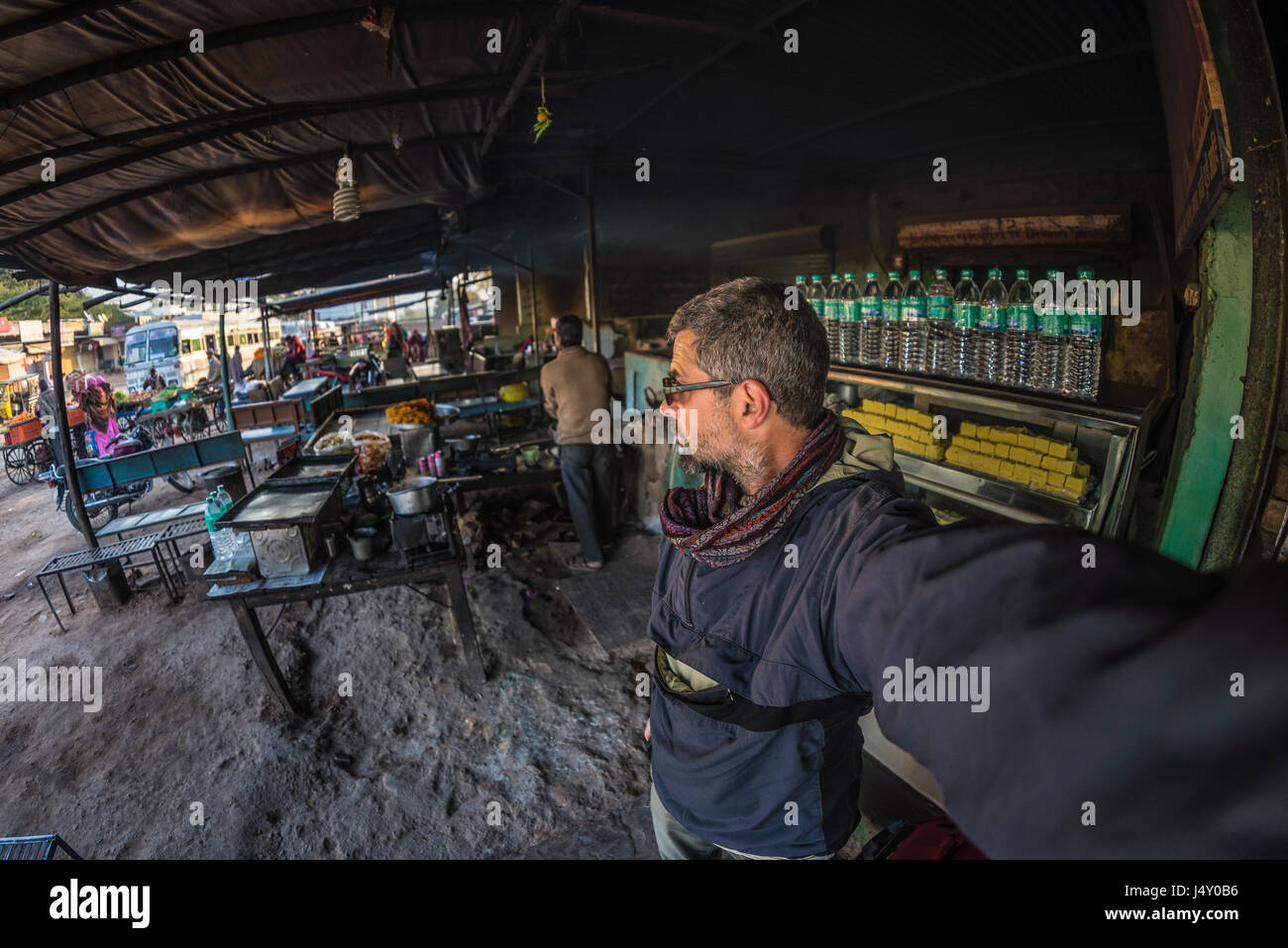 Tourist taking selfie at street food stall in India bus station while traveling. Fish eye view with indian sweets in foreground. Stock Photo