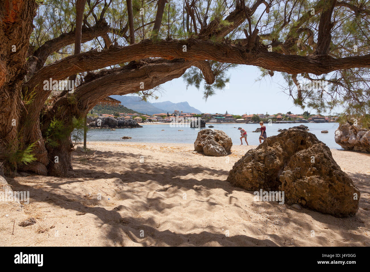 mother and daughter pass rocks and old tree on sandy stoupa beach on greek peninsula mani on peloponnese in spring with sea and old town in the backgr Stock Photo