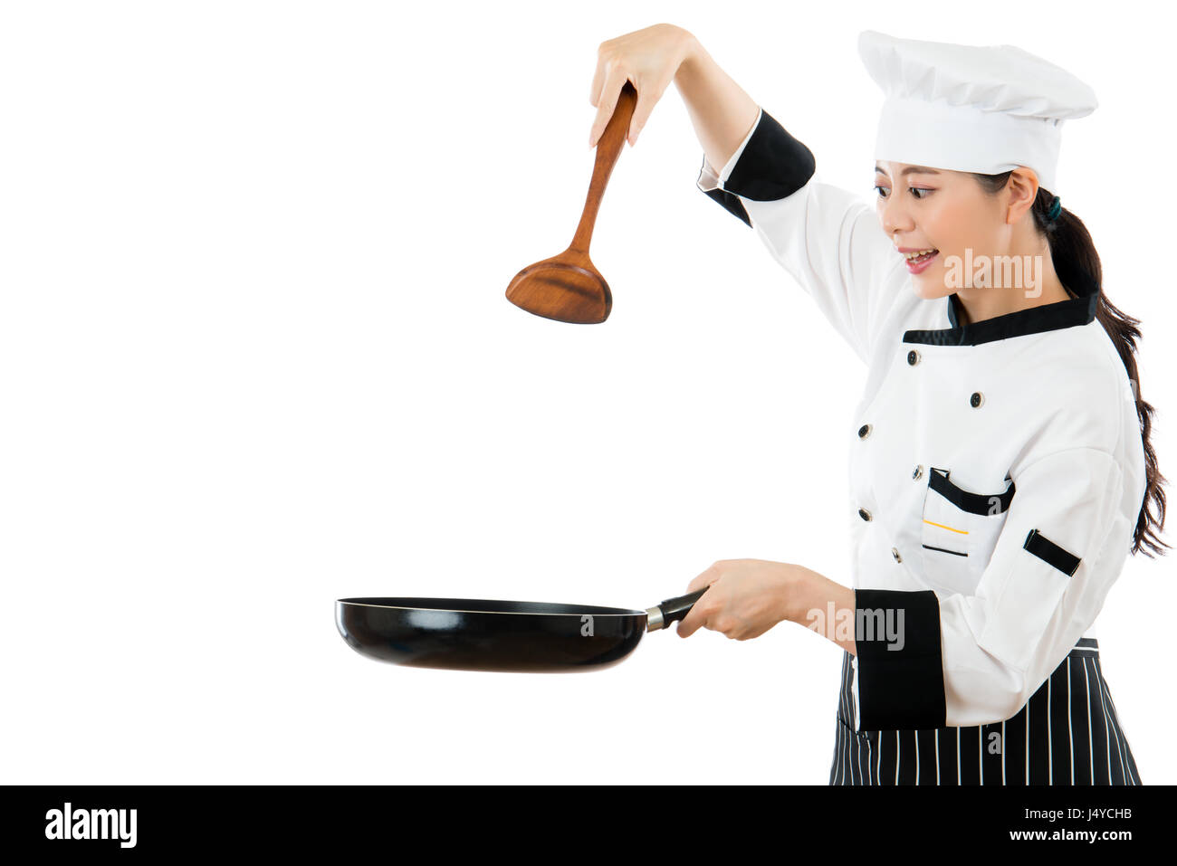 beautiful chef holding a wooden spatula in the air showing cooking gesture with copyspace and holding a pan smile standing on a white wall background. Stock Photo
