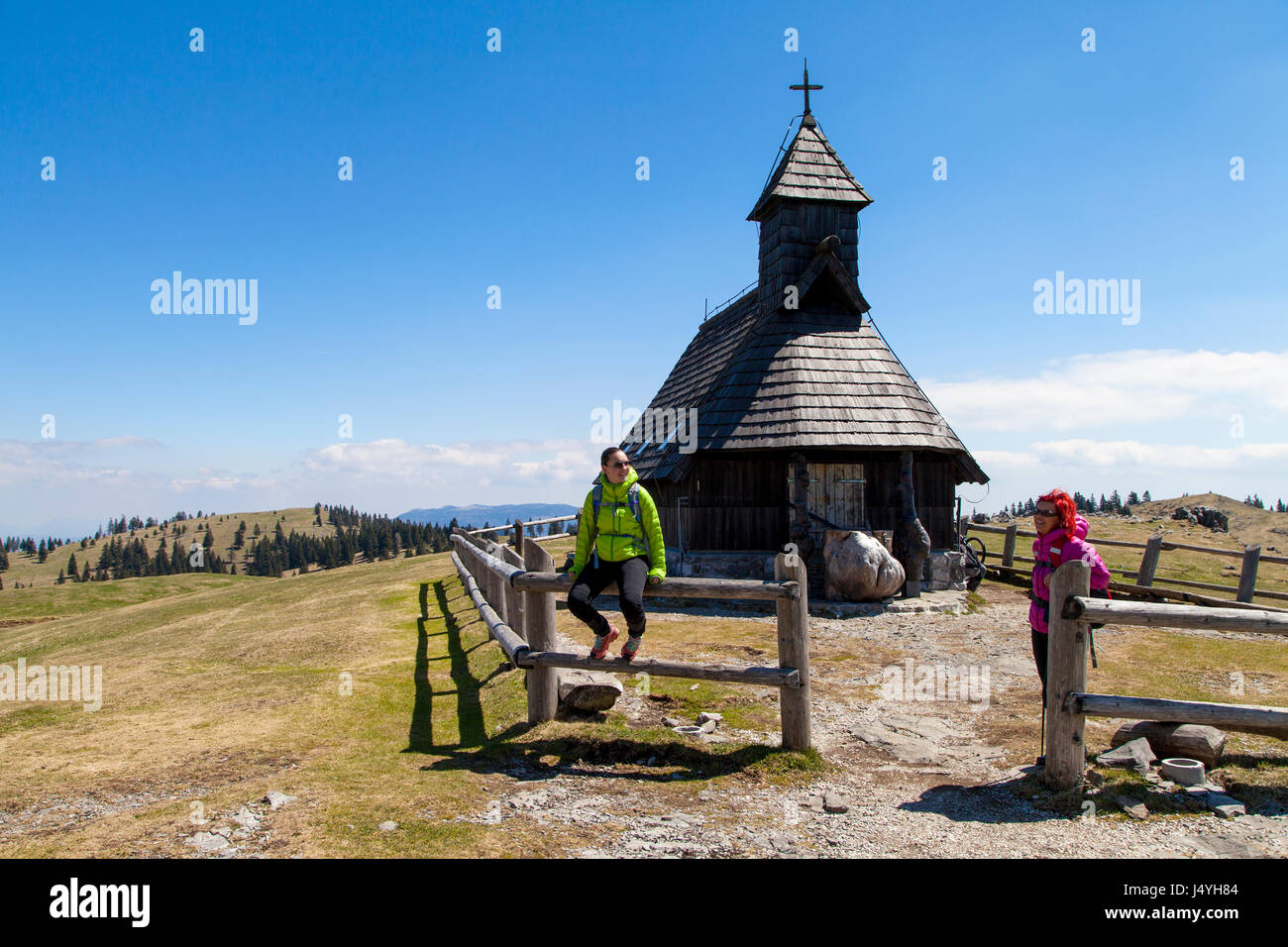 Happy smiling hikers resting near small mountain church in mountain nature on sunny day, Velika planina, Slovenia, against  bright blue sky Stock Photo