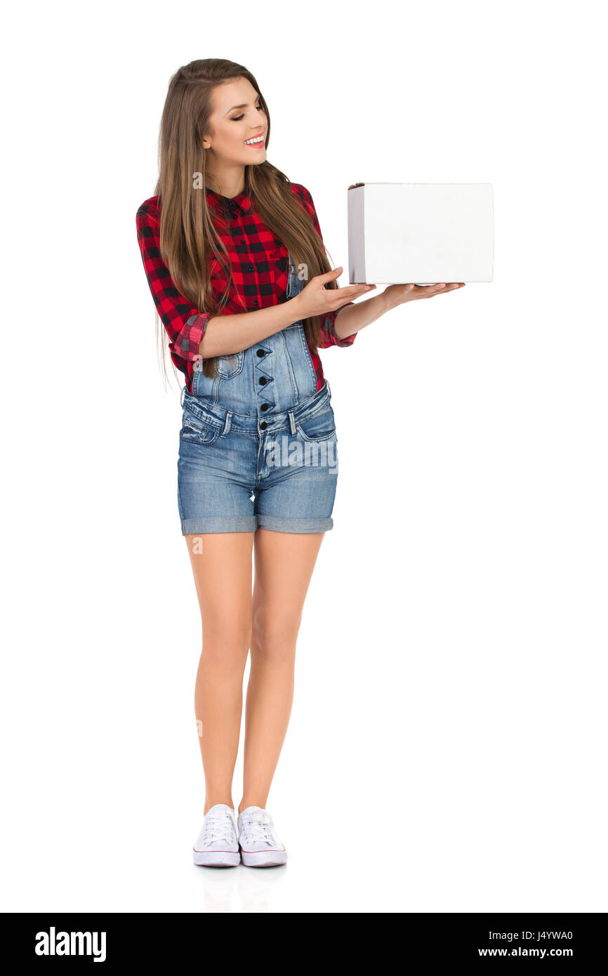 Smiling young woman in red lumberjack shirt and jeans dungarees shorts  presenting white cardboard box and looking at it. Full length studio shot  isola Stock Photo - Alamy