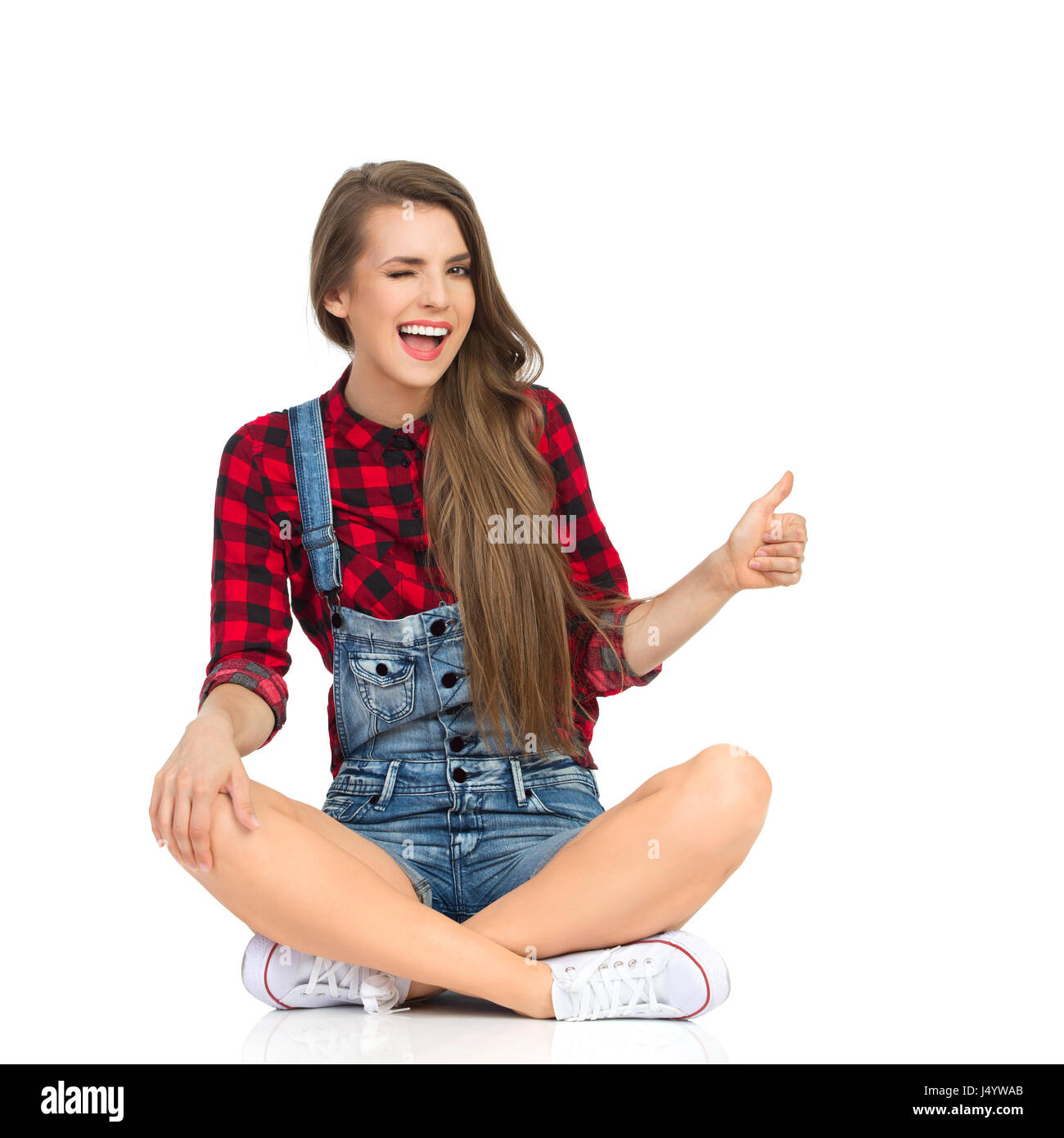Smiling girl in red lumberjack shirt, jeans dungarees shorts and white  sneakers sitting with legs crossed, showing thumbs up and winks eye, Full  lengt Stock Photo - Alamy