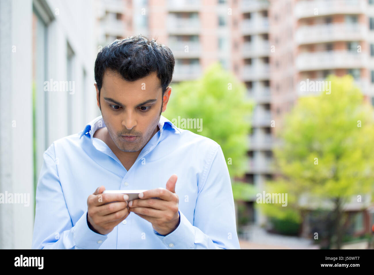 Closeup portrait of guy in blue shirt seeing stunning text messages on cell smart phone, isolated city outside background Stock Photo