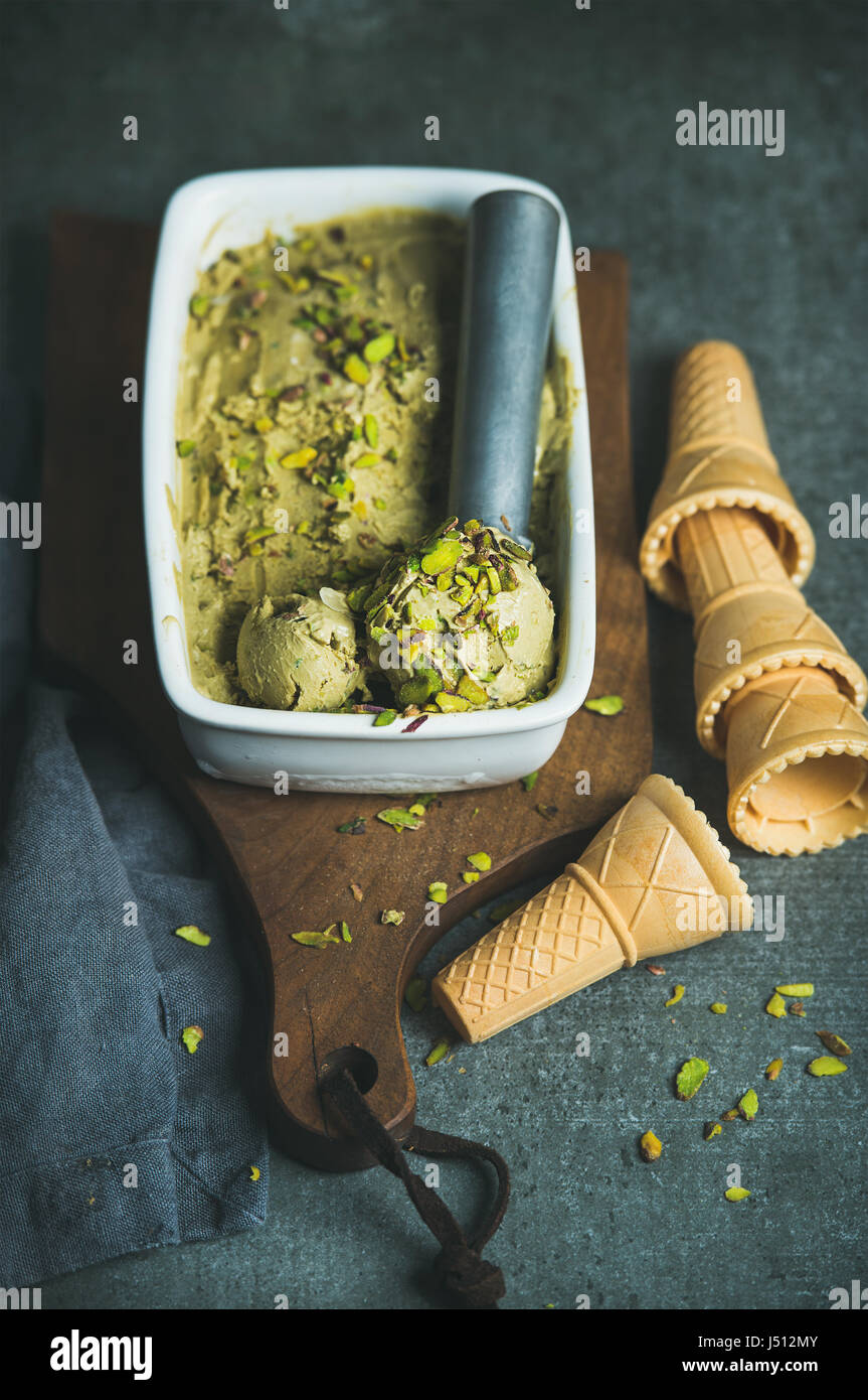Homemade pistachio ice cream in ceramic mold with metal scooper, crashed pistachio nuts and waffle cones over dark concrete background, selective focu Stock Photo