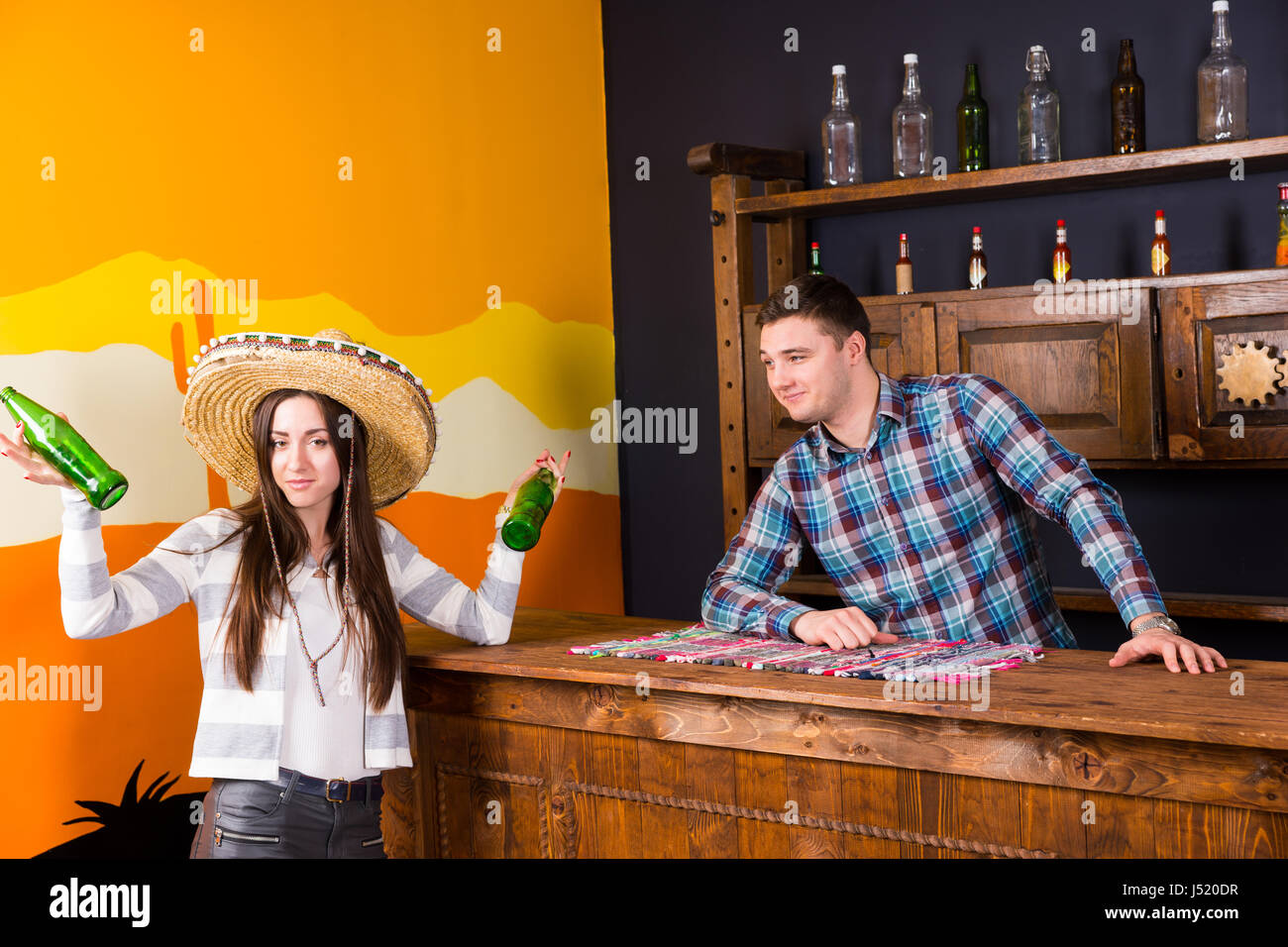 A young woman in a sombrero holding two bottles of beer and standing at the bar next to the bartender in a plaid shirt in a Mexican bar Stock Photo