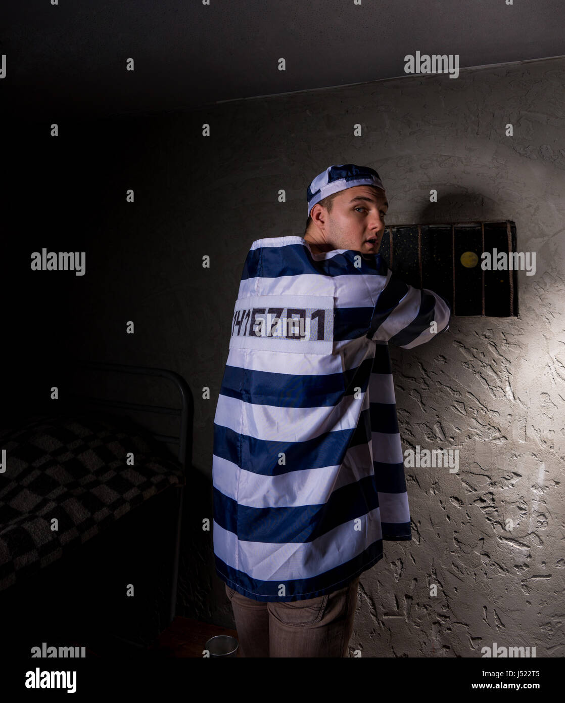 Prisoner wearing prison uniform looked back while trying to escape through the window with bars in a small dark prison cell Stock Photo