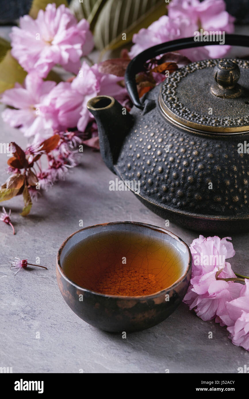 Black iron teapot and traditional ceramic cup of tea with blossom pink flowers cherry branch over gray texture background. Asian style. Stock Photo