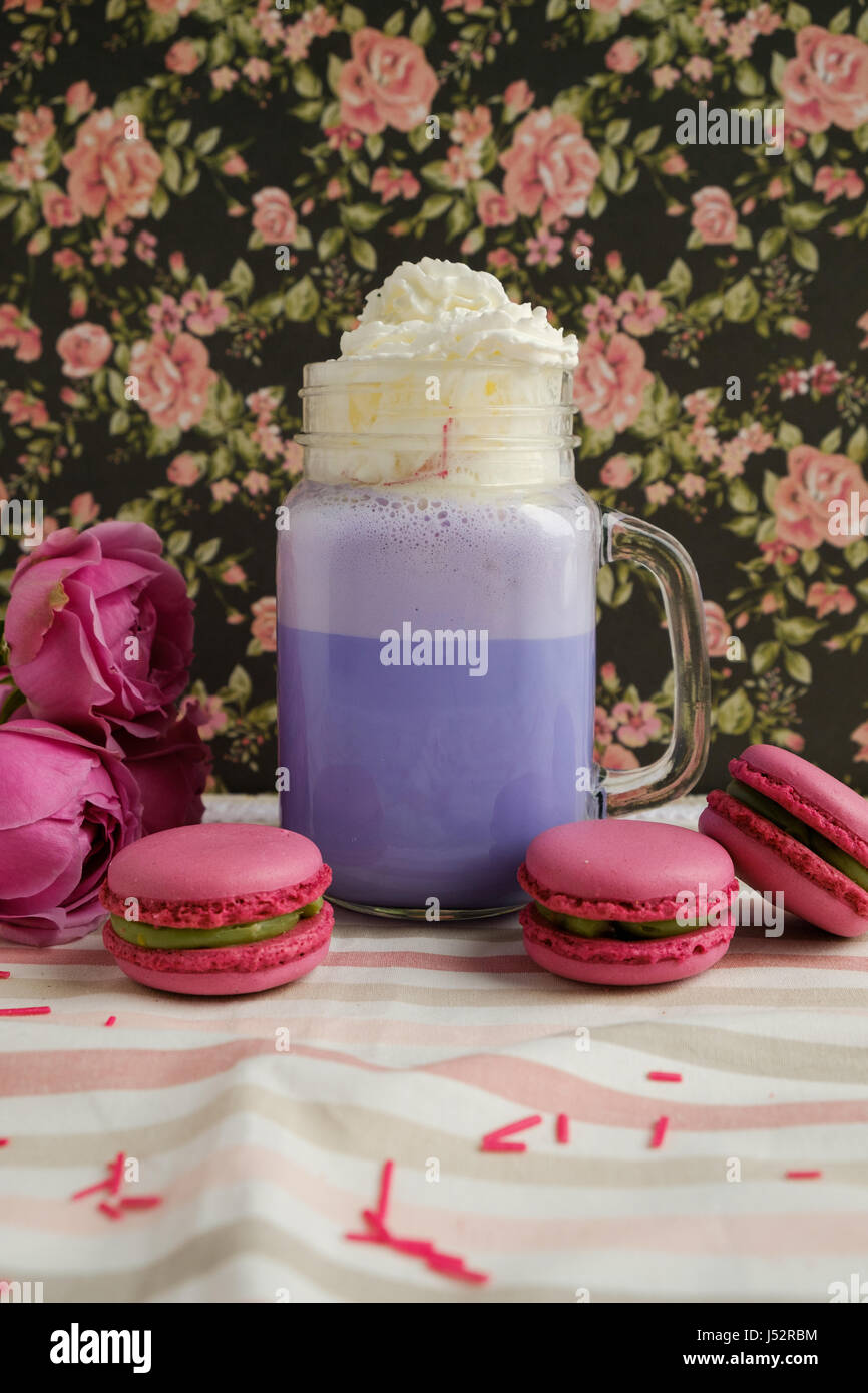 Purple coffee in stylized mason jar cup with macarons and roses and colorful decoration on background of floral pattern. Blueberry milk shake. Unicorn Stock Photo