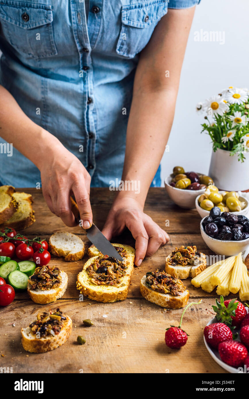 A woman spreading olive tapenade on a slice of corn bread photographed from front view. Three types of olives, pickled baby corns, strawberries, tomat Stock Photo