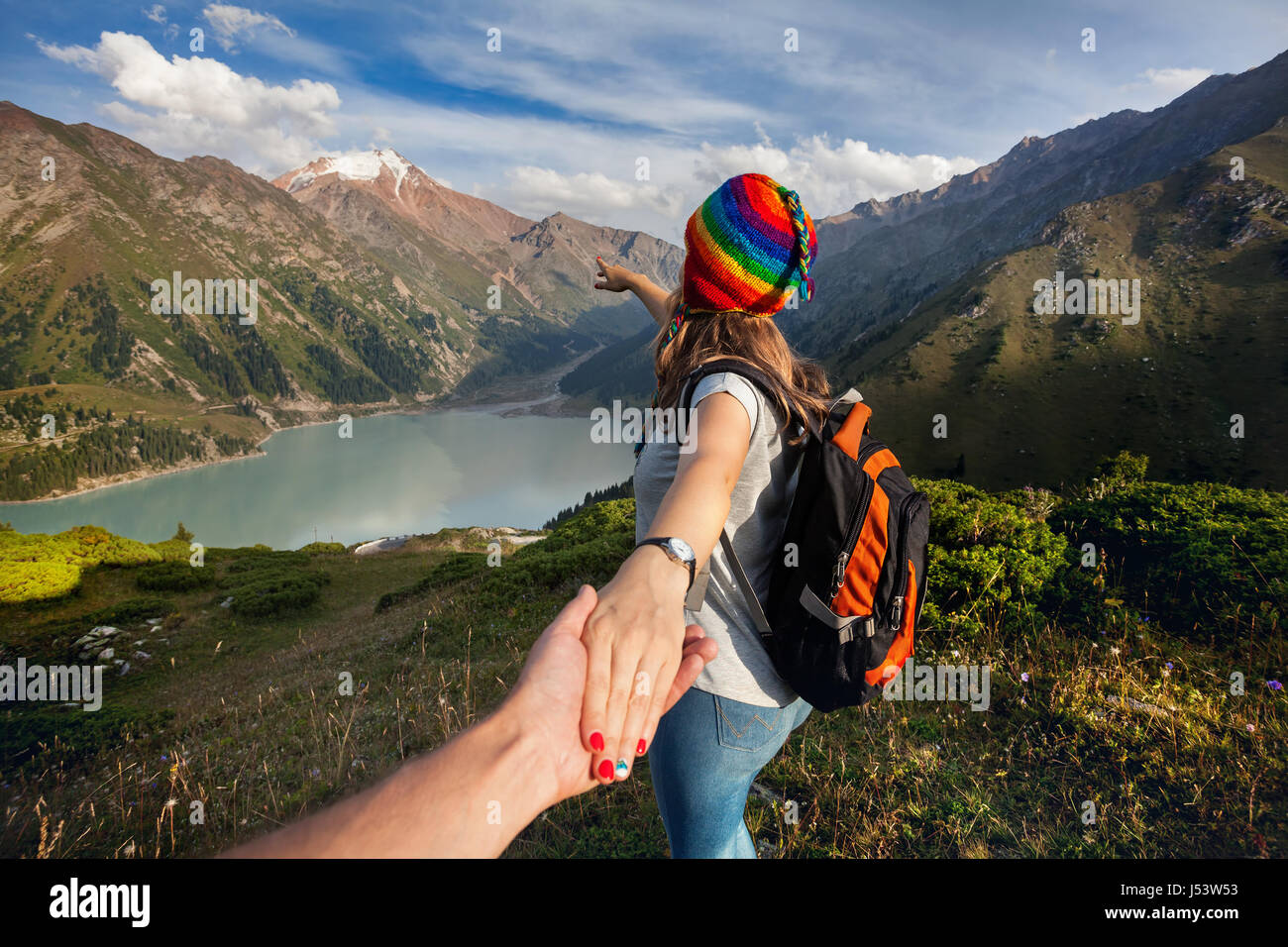 Tourist woman in rainbow hat and brown poncho holding man by hand and pointing to the lake in the mountains Stock Photo