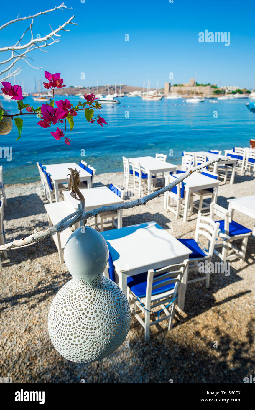 Simple beachside tables line the pebble shore of the tourist resort of Bodrum, Turkey with a scenic view of the castle Stock Photo