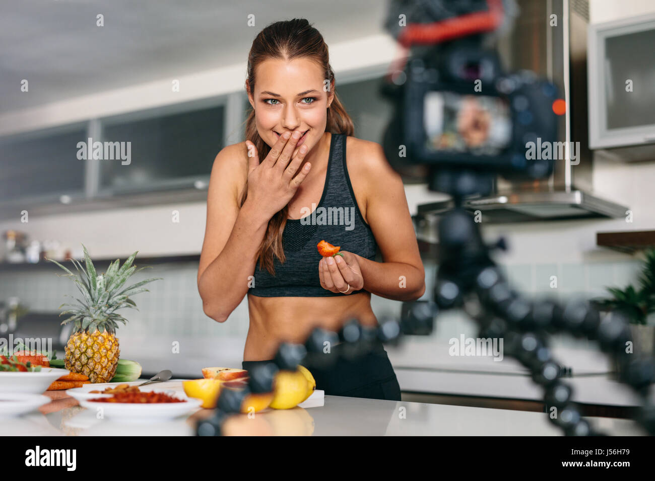 Young lady eating a strawberry at the kitchen table facing the camera. Smiling woman recording content for her vlog in kitchen. Stock Photo