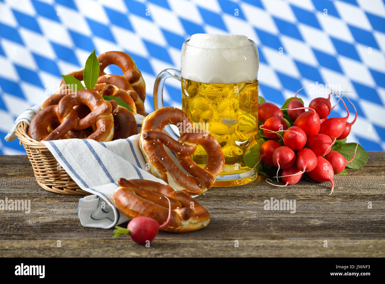 Fresh pretzels in a breadbasket, a bunch of radishes and a Bavarian beer mug on a wooden table, in the background the white-blue flag of Bavaria Stock Photo