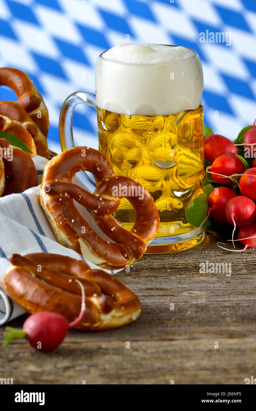 Fresh pretzels in a breadbasket, a bunch of radishes and a liter of Bavarian beer on a wooden table, in the background the white-blue flag of Bavaria Stock Photo