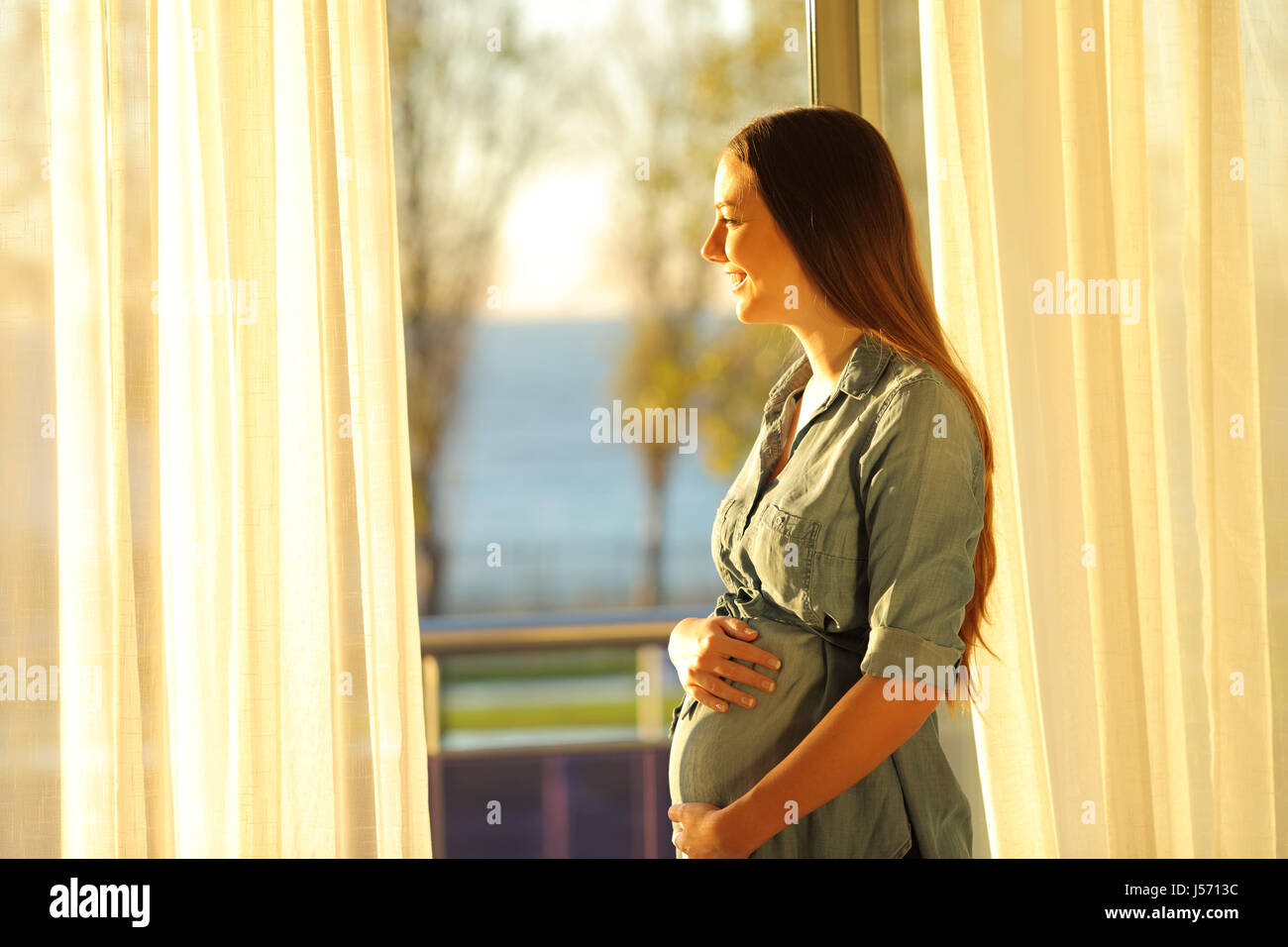 Side view of a pregnant woman looking outdoors through a window at sunset with a warmth light Stock Photo