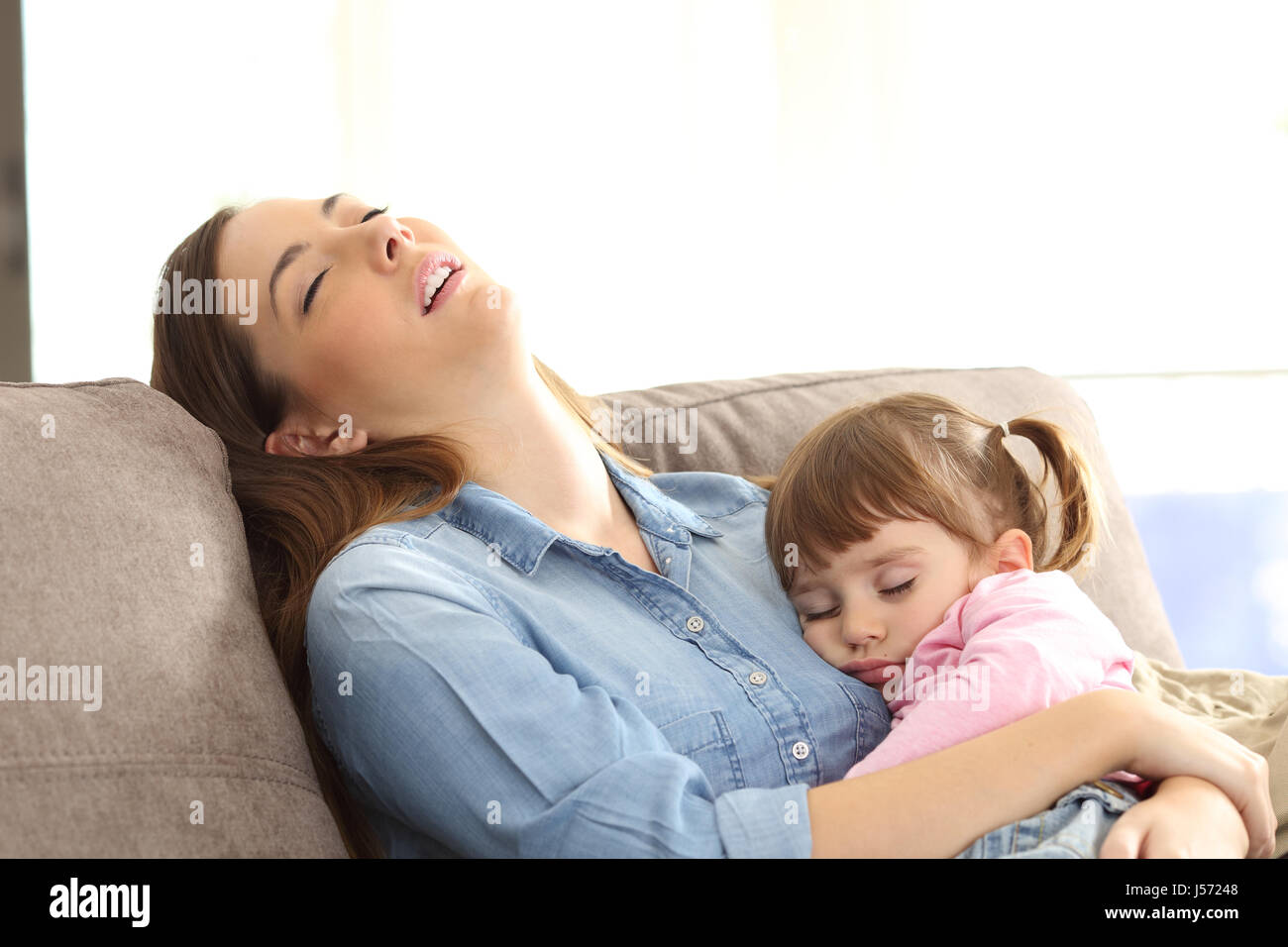 Tired mother sleeping embracing to her asleep baby daughter sitting on a sofa at home Stock Photo