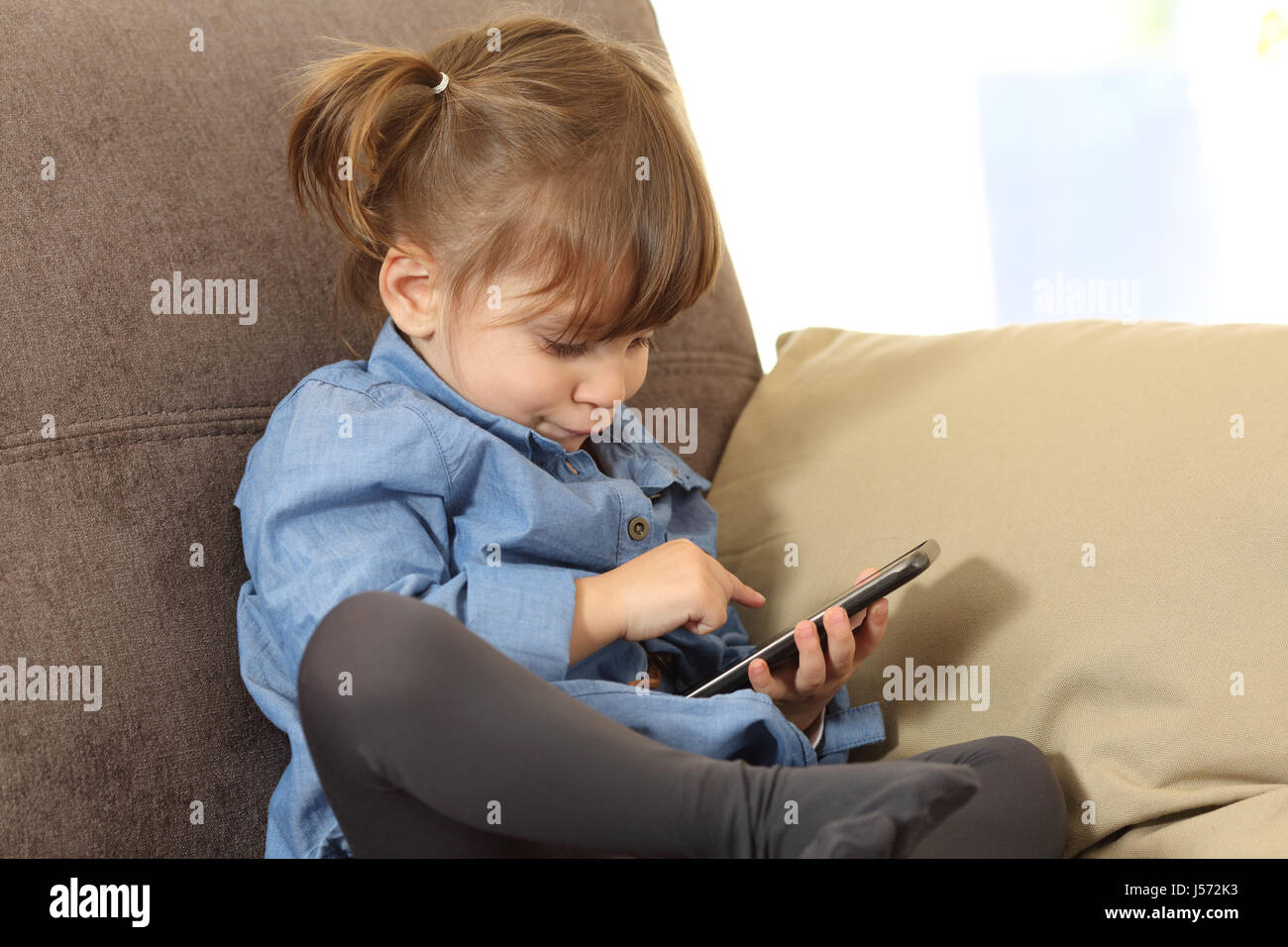 Concentrated baby playing games with a smart phone on line sitting on a sofa in the living room at home Stock Photo