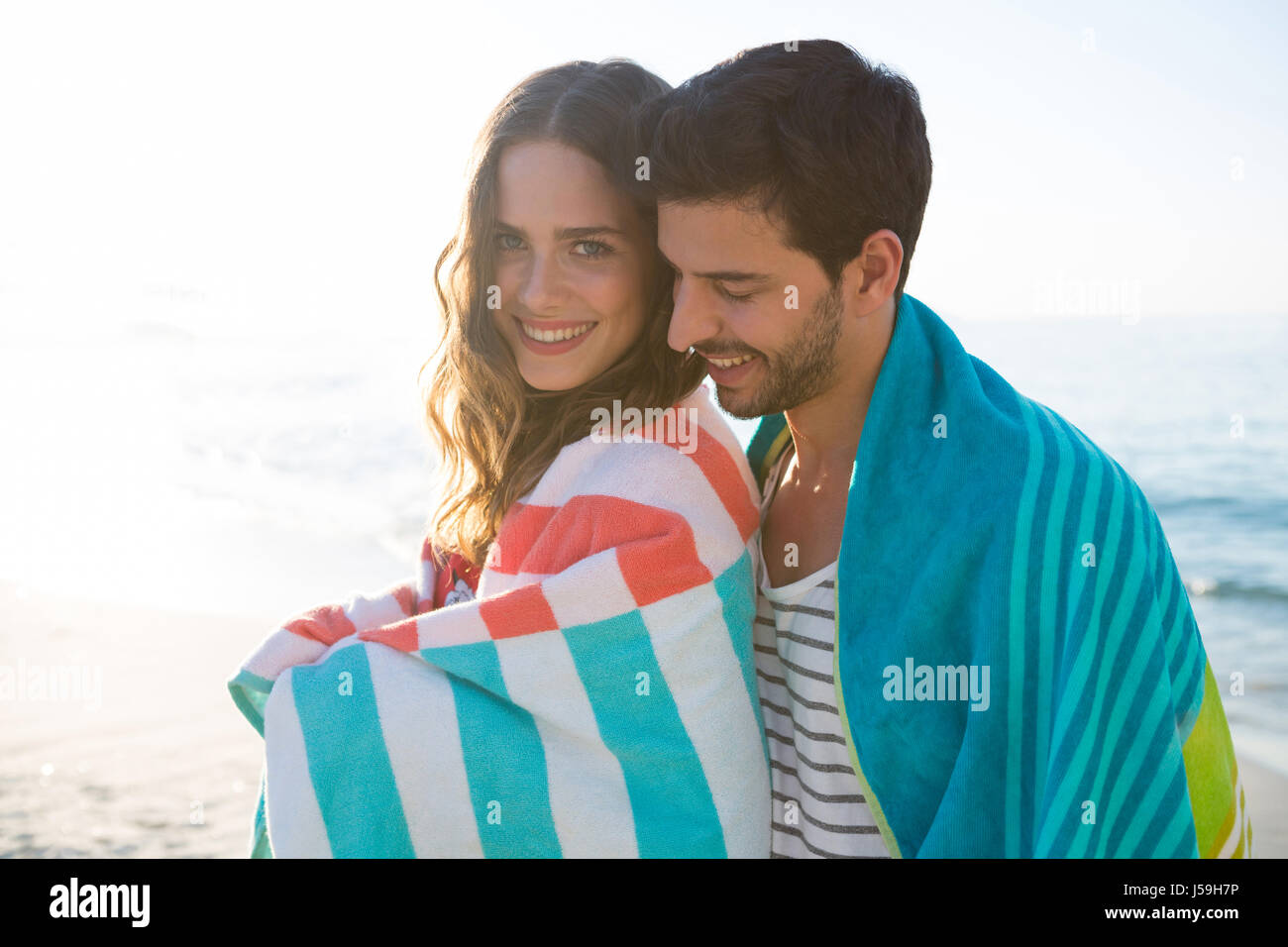 Happy young couple wrapped in blanket at beach on sunny day Stock Photo ...