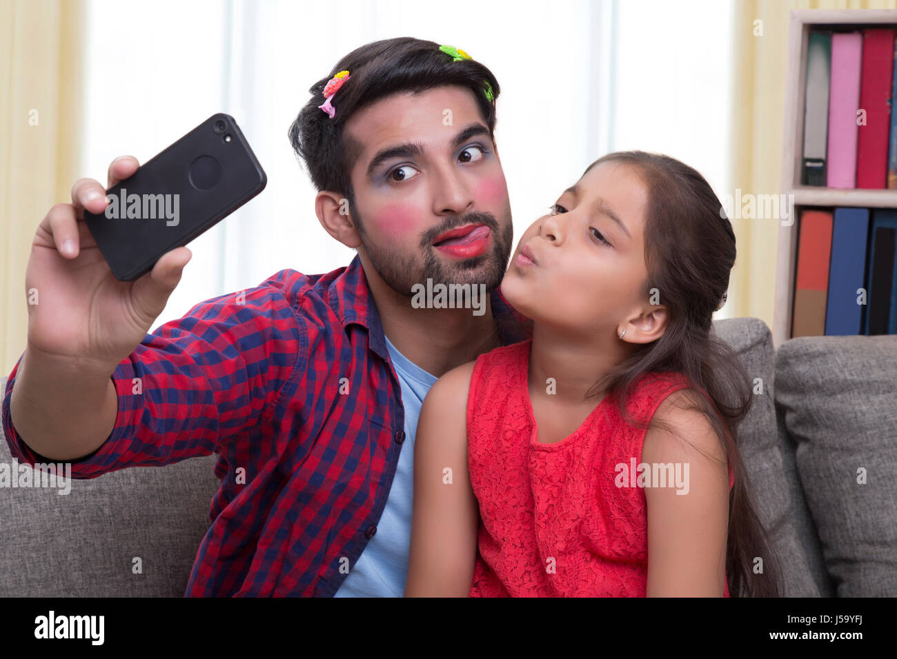 Father wearing make-up pulling funny faces sitting with daughter taking selfie Stock Photo