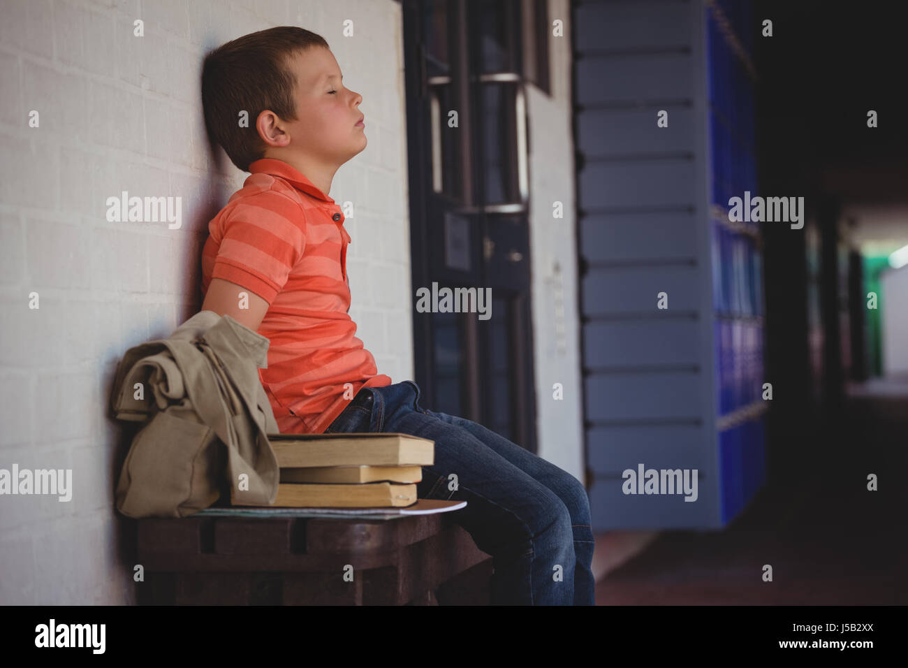 Sad boy with eyes closed sitting on bench by wall in corridor at school Stock Photo