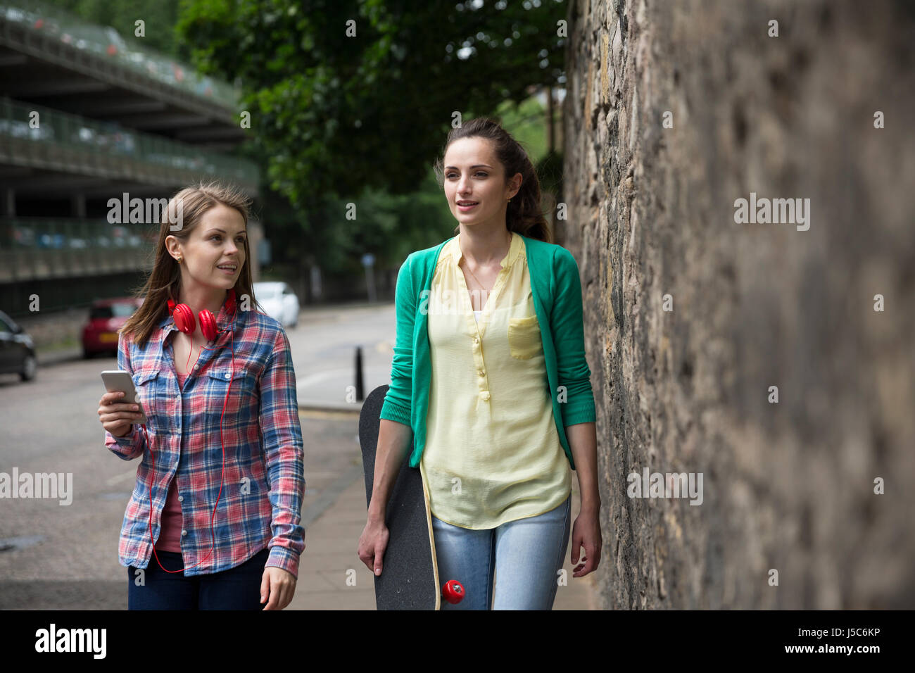 Two fashionable women walking down a city street, one with a skateboard. Stock Photo